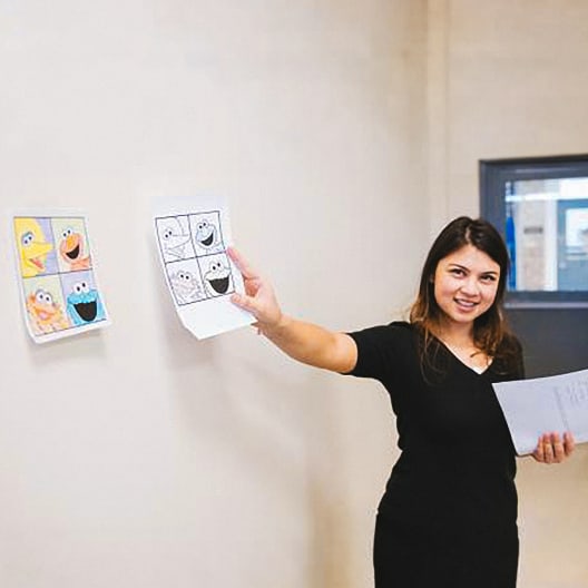 A woman stands by a wall, smiling and holding up printed sheets with cartoon faces similar to Sesame Street characters. Another sheet with similar cartoon faces is taped to the wall beside her.