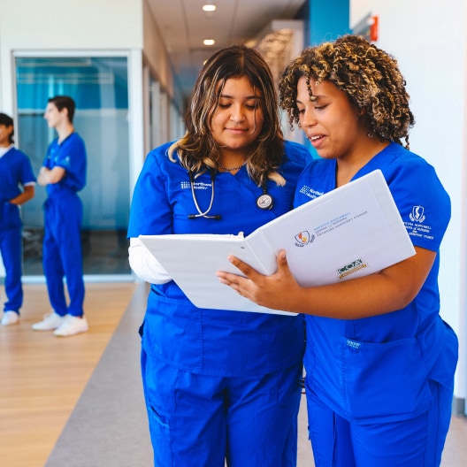 Two medical students in blue scrubs review a file together in a hallway, while two other students stand in the background talking.
