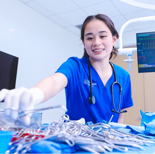A medical professional in blue scrubs and gloves smiles while reaching for a surgical instrument on a tray in a brightly lit hospital room.
