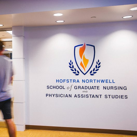 A person walks past a wall with the Hofstra Northwell School of Graduate Nursing and Physician Assistant Studies logo and name displayed in blue and orange.