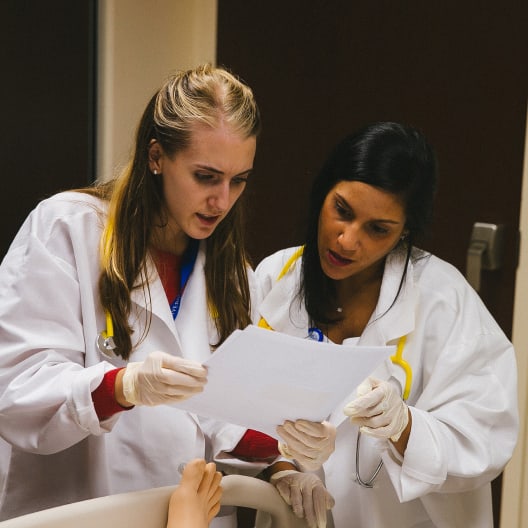 Two healthcare professionals in white coats and gloves review a document together while standing next to a patient’s bed, focusing intently on the paper. A patients foot is visible in the foreground.