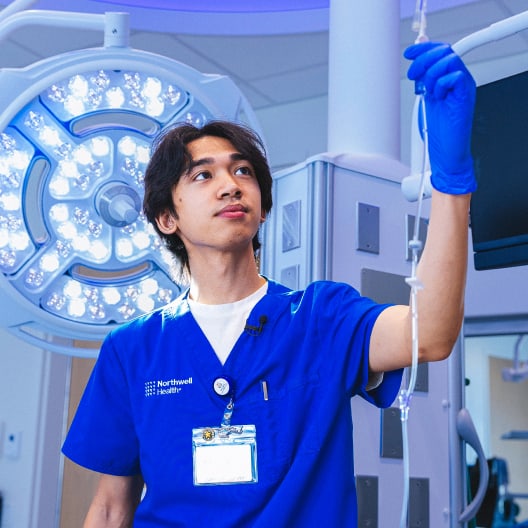 A healthcare professional in blue scrubs and gloves adjusts an IV drip in a brightly lit hospital room, with surgical lights and medical equipment visible in the background.