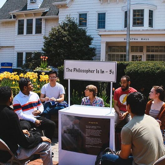 A group of people sit in a circle outdoors, engaging in conversation around a booth with a sign reading The Philosopher Is In – 5¢ in front of a building labeled Hofstra Hall. Yellow flowers bloom nearby.
