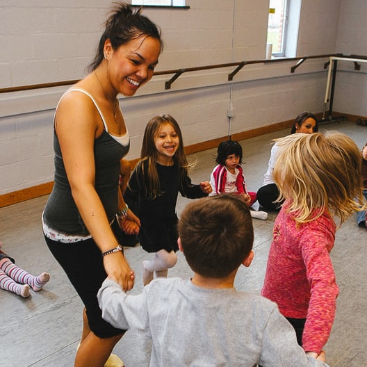 A woman leads a group of young children in a circle dance in a brightly lit studio with ballet barres along the wall. The children are smiling and appear to be having fun.