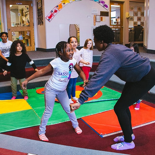 A group of young children participate in an interactive activity with an adult in a colorful classroom. The children and adult are holding hands and moving energetically on brightly colored mats.