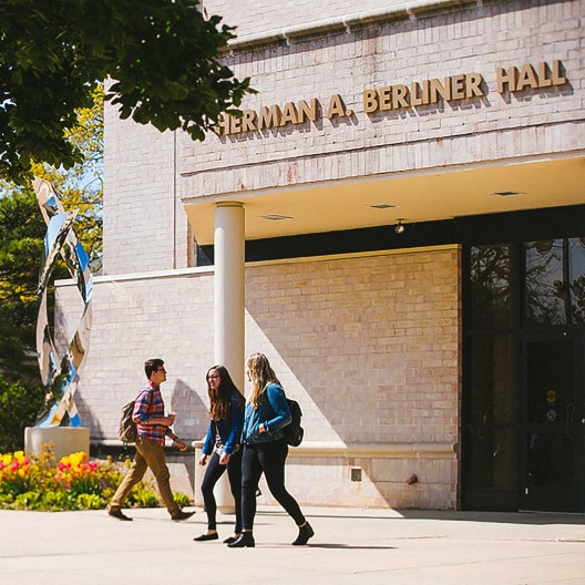 Three students walking together on a sunlit campus sidewalk in front of a building labeled “Herman A. Berliner Hall”