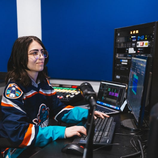 A person wearing glasses and a hockey jersey sits at a desk, using a computer and audio equipment in a studio with blue walls and multiple screens.