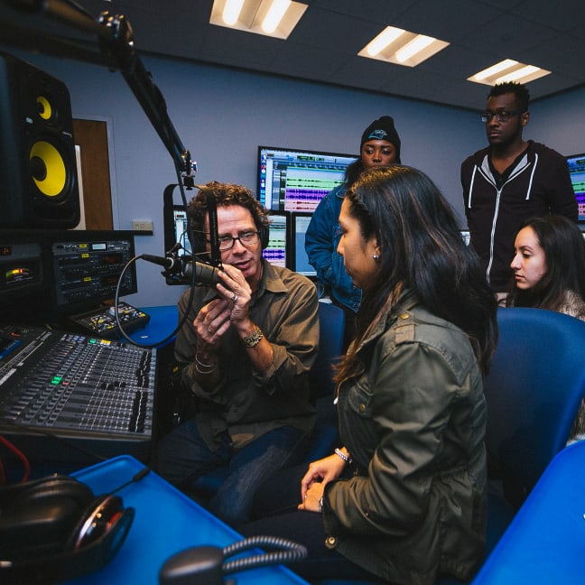 A man shows a group of four young adults how to use equipment in a recording studio, surrounded by audio consoles, microphones, and computer screens.