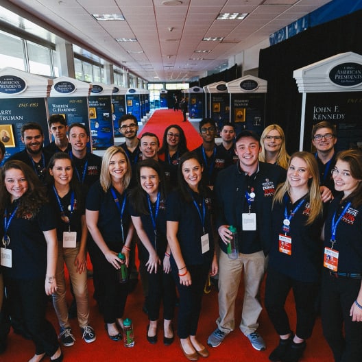 A group of young adults wearing matching navy blue shirts and name badges stand together, smiling, in a hallway with a red carpet and exhibit displays in the background.