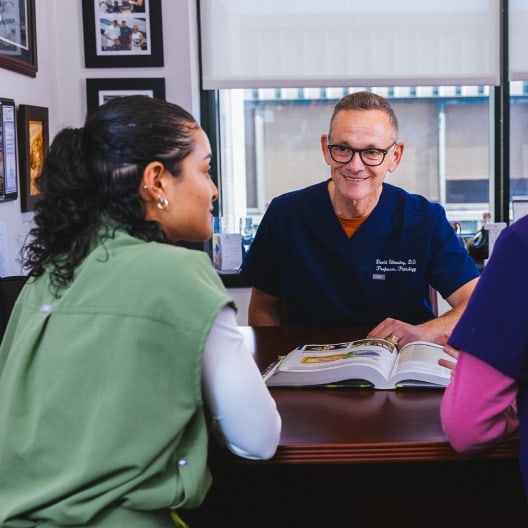 Three people sit around a desk in an office. One man in blue scrubs is smiling and speaking to two women, one in green scrubs and the other in purple. An open book is on the desk. Framed photos hang on the wall.