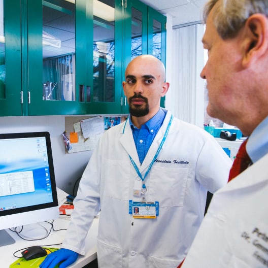 Two scientists in white lab coats talk in a laboratory. One stands by a computer with laboratory equipment nearby, while the other faces him. Green cabinets and lab materials are visible in the background.