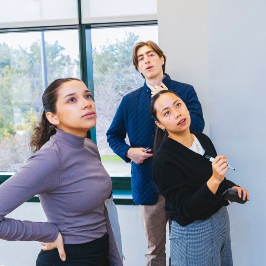 Three people stand in front of a whiteboard, appearing focused and thoughtful. One woman holds a marker, ready to write, while the others look at the board. A window with trees outside is in the background.