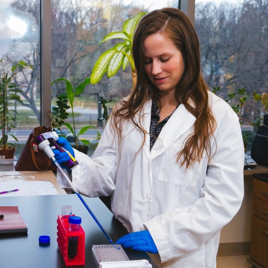A woman in a white lab coat and blue gloves uses a pipette to transfer liquid into a tray in a laboratory setting, with plants and trees visible outside the window.