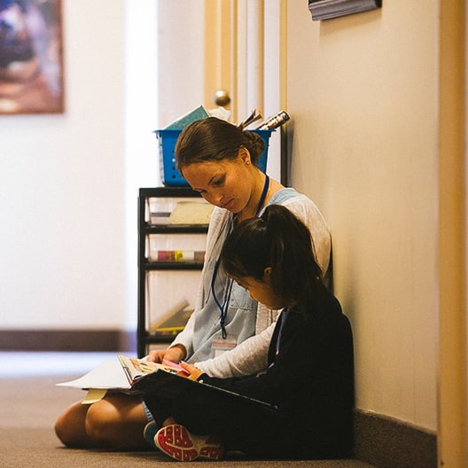 A woman and a young girl sit on the floor in a hallway, leaning against the wall as the woman helps the girl read a book. Shelves with books and supplies are visible behind them.