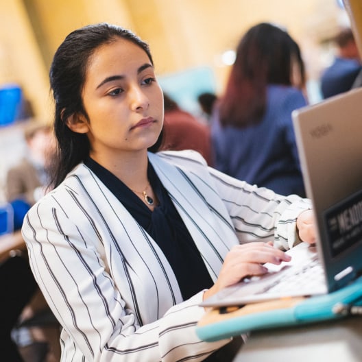 A young woman in a striped blazer works on a laptop at a table in a busy, brightly lit café or shared workspace with other people in the background.