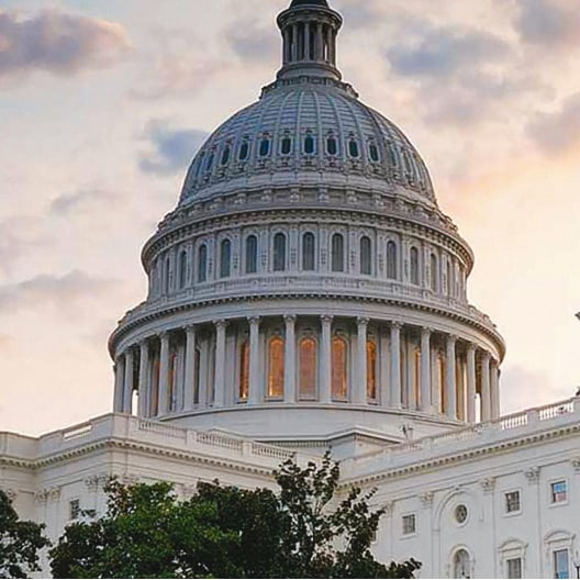 The United States Capitol building is shown at sunset, with its large dome and white columns visible against a partly cloudy sky. Trees are in the foreground.