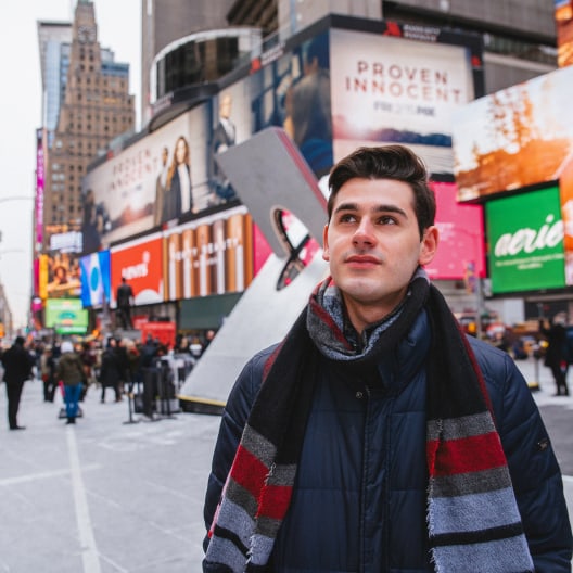 A young man with dark hair and a striped scarf stands in a busy city square surrounded by colorful billboards and tall buildings, looking upward with a slight smile. People walk in the background.