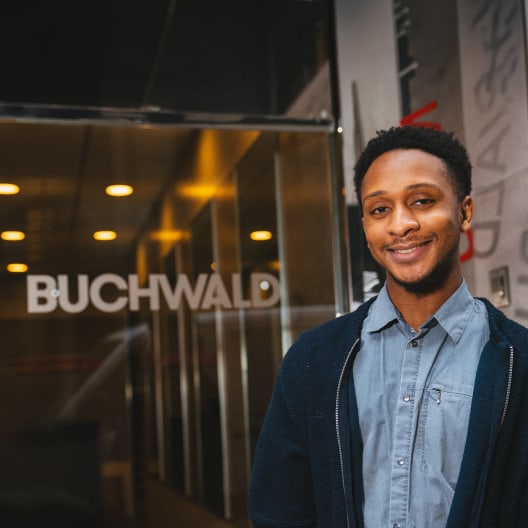 A young man in a blue shirt and jacket smiles while standing in front of a glass entrance displaying the word BUCHWALD. The setting appears to be indoors, with warm lighting and modern decor.