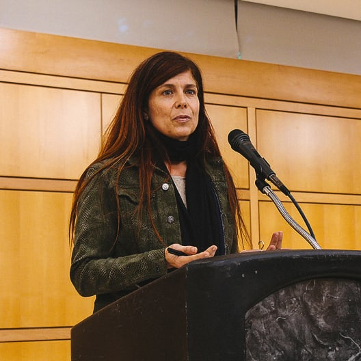 A woman with long brown hair stands at a podium, speaking into a microphone. She is wearing a green jacket and black scarf, with a wooden paneled wall in the background.