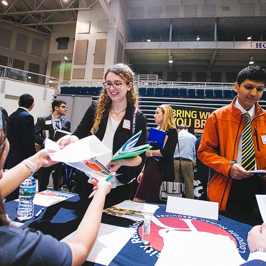 A young woman smiles while handing papers across a table at a busy indoor career fair. Other people stand nearby, holding folders and talking with recruiters under bright overhead lights.
