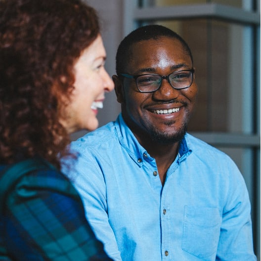 Two people indoors, one in focus smiling at the camera wearing glasses and a light blue shirt, and another person in the foreground out of focus, both appearing to enjoy a conversation.