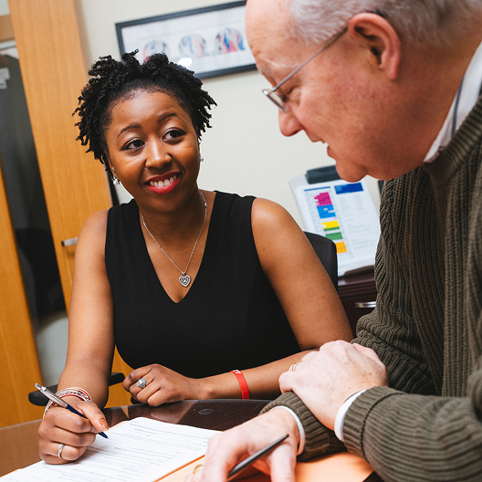 A woman smiles while holding a pen and looking at an older man across a desk. They appear to be discussing documents in an office setting.