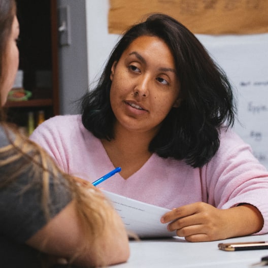 A woman with dark hair wearing a pink sweater sits at a table, holding a pen and paper, and appears to be attentively listening to another person during a conversation.