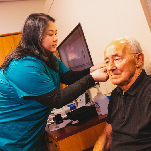 A female healthcare worker in teal scrubs examines the ear of an elderly man seated in a clinic room. Medical tools and supplies are visible on the counter beside them.