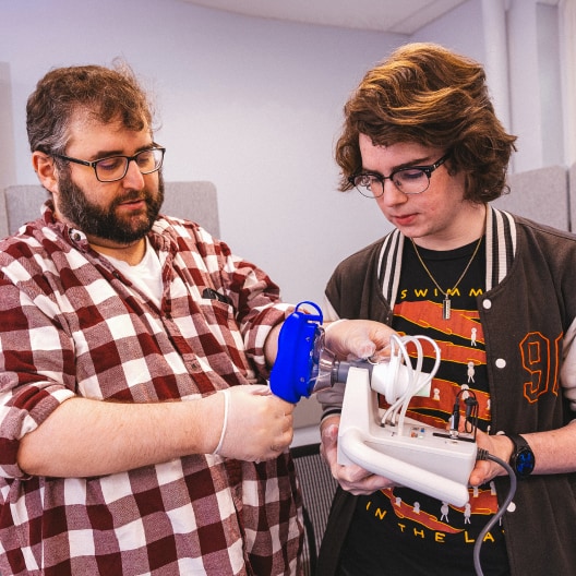 Two people wearing glasses examine a mechanical device with wires and a blue component. One person wears gloves and a red plaid shirt; the other wears a T-shirt and jacket. They appear to be in a lab or classroom setting.