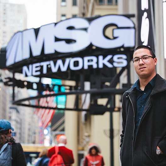 A man wearing glasses and a black coat stands outside near a large MSG Networks sign in an urban setting with tall buildings in the background.