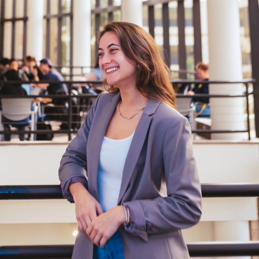 A young woman with long brown hair, wearing a light gray blazer and white top, smiles while standing indoors in a modern building with large windows and people sitting in the background.