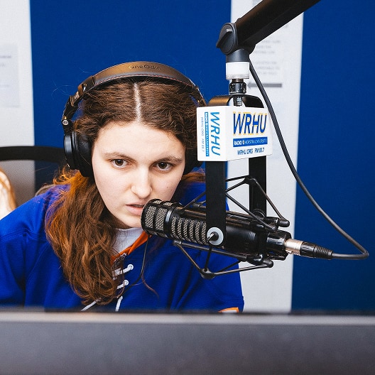 A woman with long brown hair wearing headphones speaks into a microphone at a radio station. A sign on the microphone reads WRHU. She is focused on something in front of her, with a blue and white background.