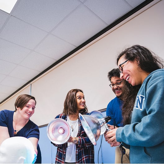 Four people stand together indoors, smiling and laughing while holding large heat lamps. They appear to be participating in a group activity or workshop in a brightly lit room with a white ceiling.