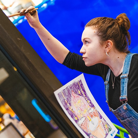 A young woman in overalls paints on a vertical surface, referencing a colorful drawing taped nearby. She has her hair tied back and is focused on her work, with a vivid blue background behind her.