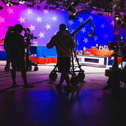 Television studio with cameramen and crew filming a brightly lit set decorated with large white stars on red, white, and blue backgrounds, where people sit at tables, suggesting a political or news broadcast.