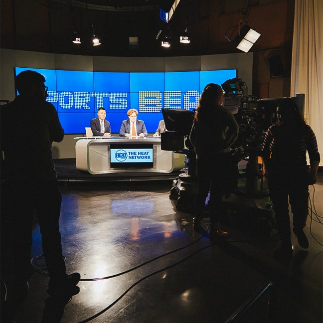TV studio scene with three people at a news desk labeled The Heat Network, brightly lit, and camera crew working in the foreground with part of the blue Sports Beat sign visible behind the hosts.