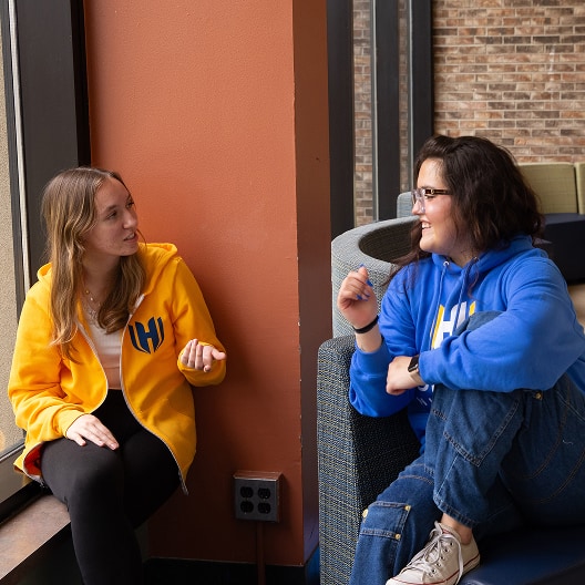 Two people wearing yellow and blue hoodies sit near a window and chat. One sits on a windowsill while the other sits on a chair, both smiling and gesturing as they talk in a brightly lit room.