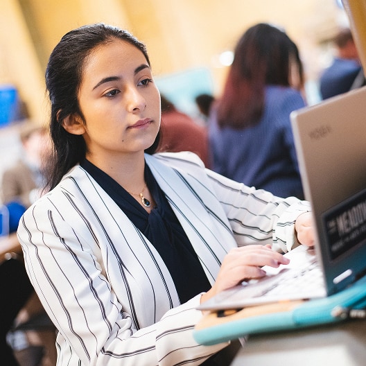 A young woman with long dark hair, wearing a white striped blazer, works on a laptop in a busy indoor setting with other people in the background.