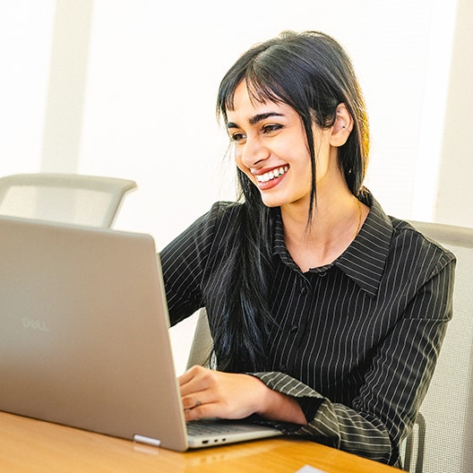 A woman with long black hair, wearing a black pinstripe shirt, smiles while working on a laptop at a desk in a bright office setting.