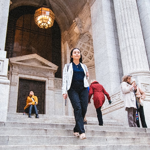 A woman walks down stone steps outside a large, ornate building with columns, as other people stand or walk nearby.