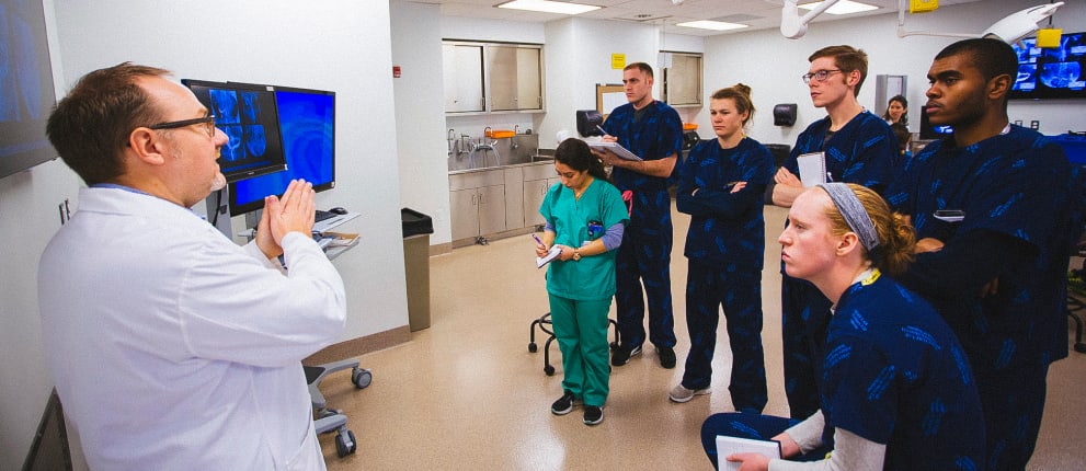 A doctor in a white coat explains something to a group of medical students in blue scrubs, who listen attentively in a hospital or clinical setting. One student in green scrubs takes notes.