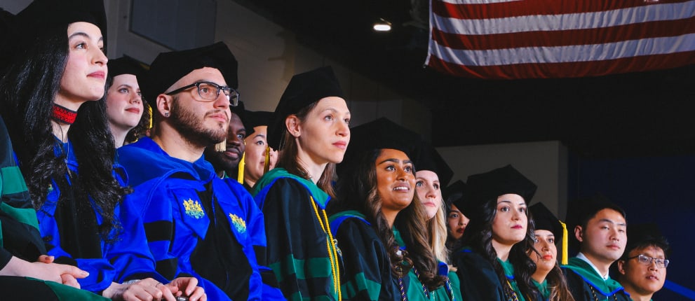 A diverse group of graduates in caps and gowns look ahead with pride and anticipation; an American flag hangs above them in the background.