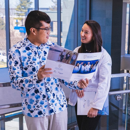 A man in a blue floral shirt holds and looks at a brochure while talking to a woman in a white coat, who is smiling. They stand indoors near large windows and a metal railing.